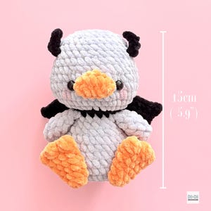 May include: A light gray crocheted plush toy with black horns and wings, an orange beak, and orange feet. The toy features black safety eyes and pink blush details. A vertical measurement line indicates a height of 15 cm or 5.9 inches.