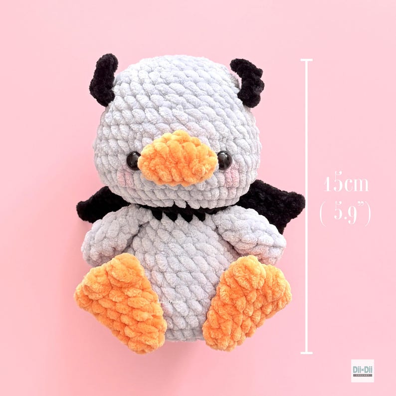 May include: A light gray crocheted plush toy with black horns and wings, an orange beak, and orange feet. The toy features black safety eyes and pink blush details. A vertical measurement line indicates a height of 15 cm or 5.9 inches.