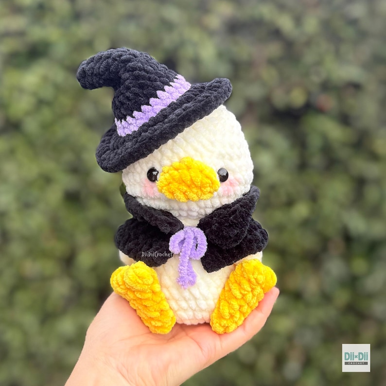 May include: A white crocheted duck plushie wearing a black witch hat with a lavender band and a black cape tied with a lavender ribbon. The duck has a bright yellow beak, yellow feet, and pink blush on its cheeks. The text Dii-Dii Crochet is visible.