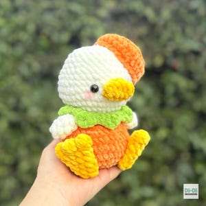 May include: A plush crochet duck toy featuring a white head with a bright orange cap, a yellow beak, and pink cheeks. It wears a green collar around its neck and has an orange body with yellow feet. The text Dii-Dii CROCHET is in the corner.