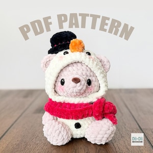 May include: A plush, light pink teddy bear wearing a snowman-themed outfit. The bear has a white hood, a black top hat with an orange pom-pom, and a red scarf. The text "PDF PATTERN" is above the bear.