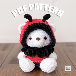 May include: A white crochet bear wearing a red and black ladybug-themed hood and collar. The bear has a brown nose and small black eyes. The words "PDF PATTERN" are displayed above the bear.