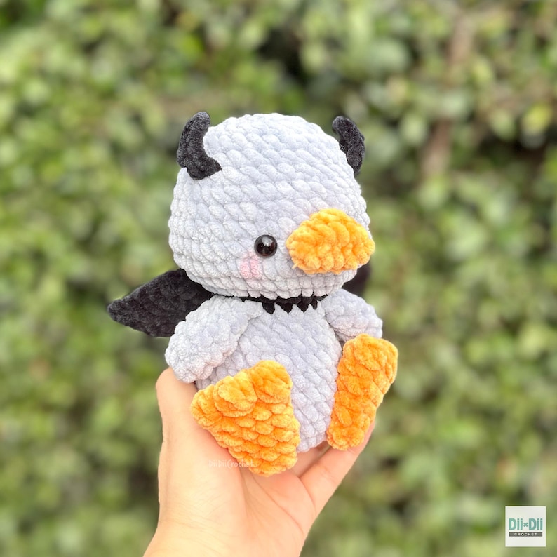 May include: A light gray crocheted plush toy shaped like a duck with black wings and small black horns on its head. It features a bright orange beak and orange webbed feet, with small pink blush marks under its black plastic eyes.