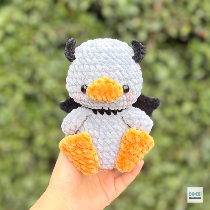 May include: A small, handmade crochet plush toy shaped like a duck with light gray yarn, bright orange feet and beak, black wings, and small black horns on its head. The toy features two black plastic eyes and pink stitched blush on its cheeks.