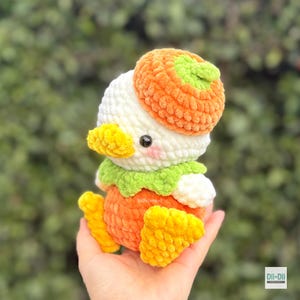 May include: A crocheted plush toy shaped like a duckling wearing an orange pumpkin hat. The duck has a white head, a yellow beak, a green collar, and an orange body with yellow feet. It features black eyes and small pink blush marks on its cheeks.