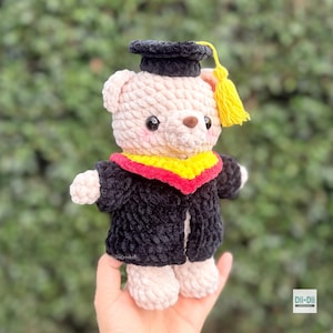 Peut inclure: Un ours en peluche crochet&eacute; portant une toque et une toge de fin d'&eacute;tudes. L'ours est rose clair avec une toque et une toge noires, un col jaune et rouge et un gland jaune. L'ours mesure environ 15 cm de haut.