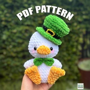 May include: A white and orange crochet duck wearing a green leprechaun hat and bow tie. The duck has a yellow beak and feet. The text "PDF PATTERN" is displayed above the duck.