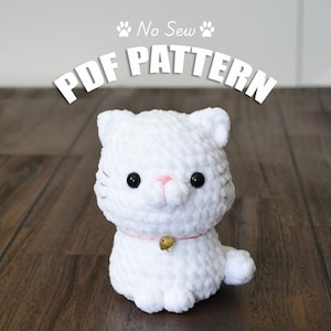 May include: White crocheted cat plush toy with black eyes, a pink nose, and a gold bell on a pink collar. The toy is sitting on a wooden surface. The text "No Sew PDF PATTERN" is above the toy.