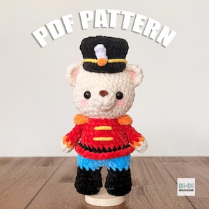 Puede incluir: Un oso de ganchillo hecho a mano vestido de soldado de juguete. El oso lleva un sombrero negro, una chaqueta roja con detalles dorados, pantalones azules y botas negras. El texto "PDF PATTERN" está arriba.