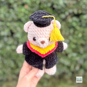 Peut inclure: Un ours en peluche crochet&eacute; portant une toque de graduation noire avec un gland jaune et une robe noire avec un col jaune et rouge. L'ours a un corps rose clair, un nez brun et de petits yeux noirs.