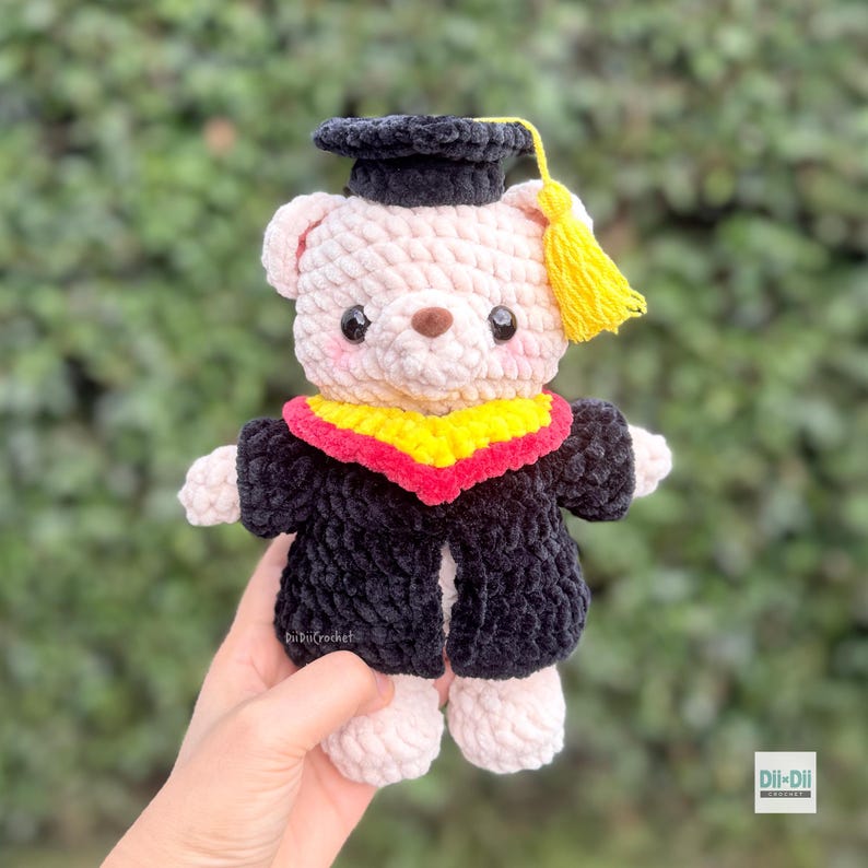Peut inclure: Un ours en peluche crochet&eacute; portant une toque et une toge de remise de dipl&ocirc;me. L'ours est rose clair avec une toque et une toge noires, un col jaune et rouge et un gland jaune. L'ours est tenu dans une main.