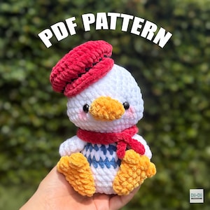 May include: A handmade crochet duckling wearing a red beret and scarf, with a yellow beak and feet. The duckling has a white body with blue and white detailing on the chest. The text "PDF PATTERN" is displayed above the duckling.
