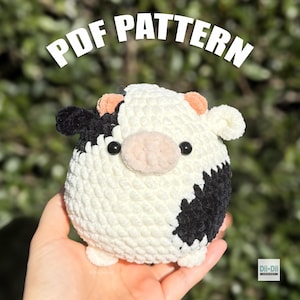 Puede incluir: Un juguete de vaca tejido a ganchillo, redondo y afelpado, hecho con hilo blanco, negro y color melocotón. La vaca tiene dos ojos negros, orejas pequeñas y un hocico color melocotón. El texto 'PDF PATTERN' aparece arriba y 'Dli-Dii CROCHET' abajo.