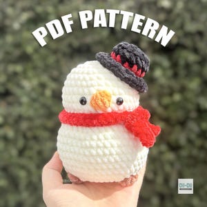 Puede incluir: Un muñeco de nieve de ganchillo blanco con un sombrero de copa negro con banda roja, una bufanda roja brillante y una nariz naranja. El texto PDF PATTERN aparece en la parte superior y un pequeño logo con Dii-Dii CROCHET en la esquina.