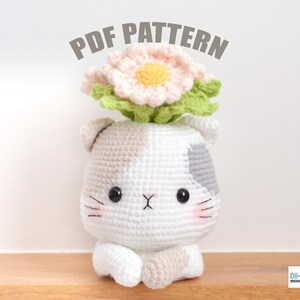 May include: Crochet pattern for a cute cat wearing a flower pot on its head. The cat is white with gray spots and has pink cheeks. The flower pot is pink with a yellow center and green leaves. The pattern is available as a PDF download.