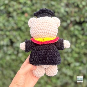Peut inclure: Un ours en crochet fait &agrave; la main portant une robe et un chapeau de graduation noirs. L'ours a un corps de couleur cr&egrave;me, un col jaune et rouge, et est tenu par une main. C'est un jouet en peluche doux.