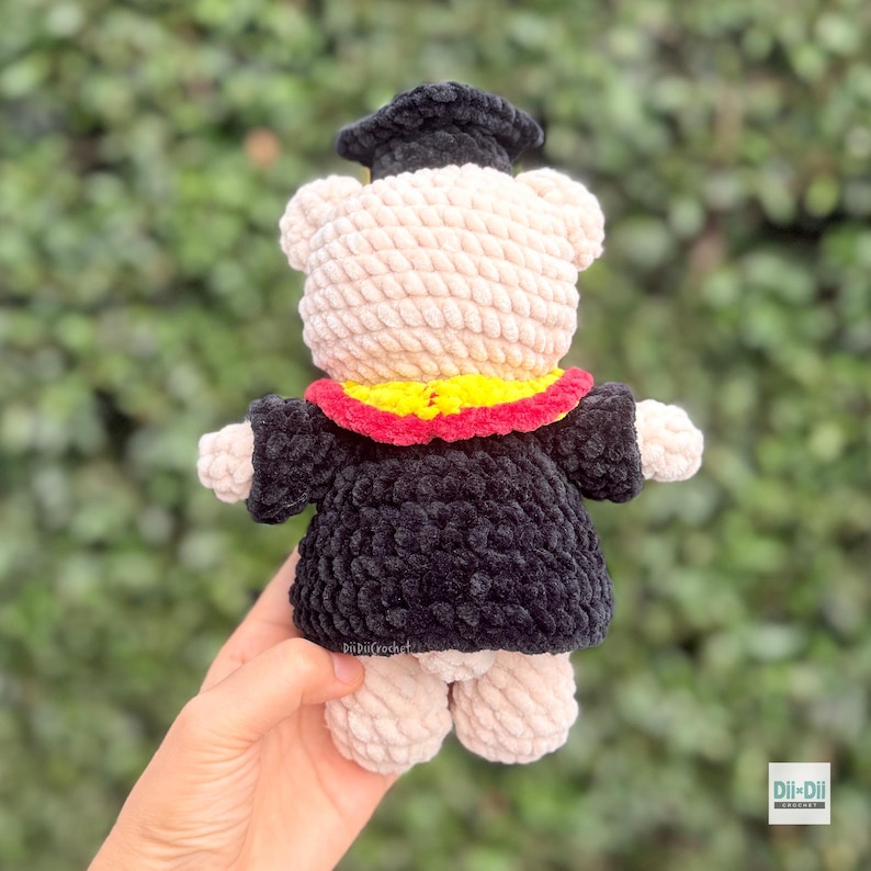 Peut inclure: Un ours en crochet fait &agrave; la main portant une robe et un chapeau de graduation noirs. L'ours a un corps de couleur cr&egrave;me, un col jaune et rouge, et est tenu par une main. C'est un jouet en peluche doux.