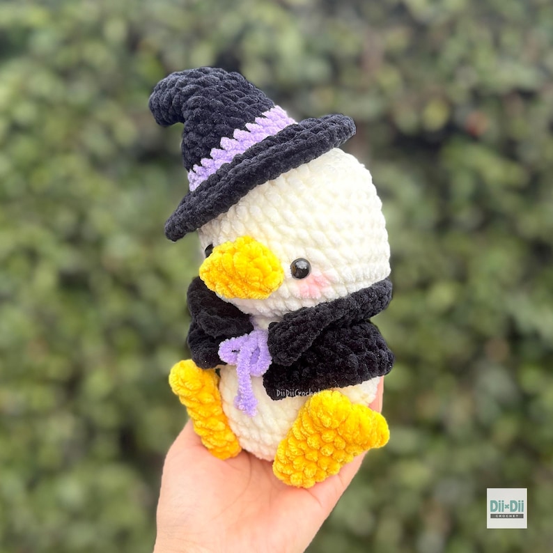 May include: A white crocheted duck plushie wearing a black witch hat with a lavender band, a black cape with a lavender tie, and bright yellow feet and beak. The plushie has small black eyes and pink blush marks on its cheeks.