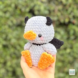 May include: A small, handmade crochet plush toy shaped like a duckling with light gray yarn, bright orange feet and beak, black wing accents, and small black eyes. The toy features a soft, textured yarn finish and a small black collar detail.