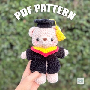 May include: A crocheted teddy bear wearing a graduation cap and gown. The bear is light pink with a black cap and gown, a yellow tassel, and a red and yellow collar. The text "PDF PATTERN" is above the bear.