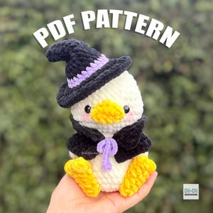 Puede incluir: Un pato de peluche tejido a ganchillo de color blanco que lleva un sombrero de bruja negro con una banda morada y una capa negra con un lazo morado. El pato tiene pico y patas amarillas, y mejillas rosadas. El texto PDF PATTERN aparece arriba.