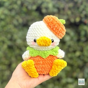 May include: A plush crochet toy shaped like a duck wearing an orange acorn cap. The duck has a white head, a yellow beak, pink cheeks, a green collar, and an orange body with yellow feet. The text Dil-Dil Crochet is visible in the bottom corner.