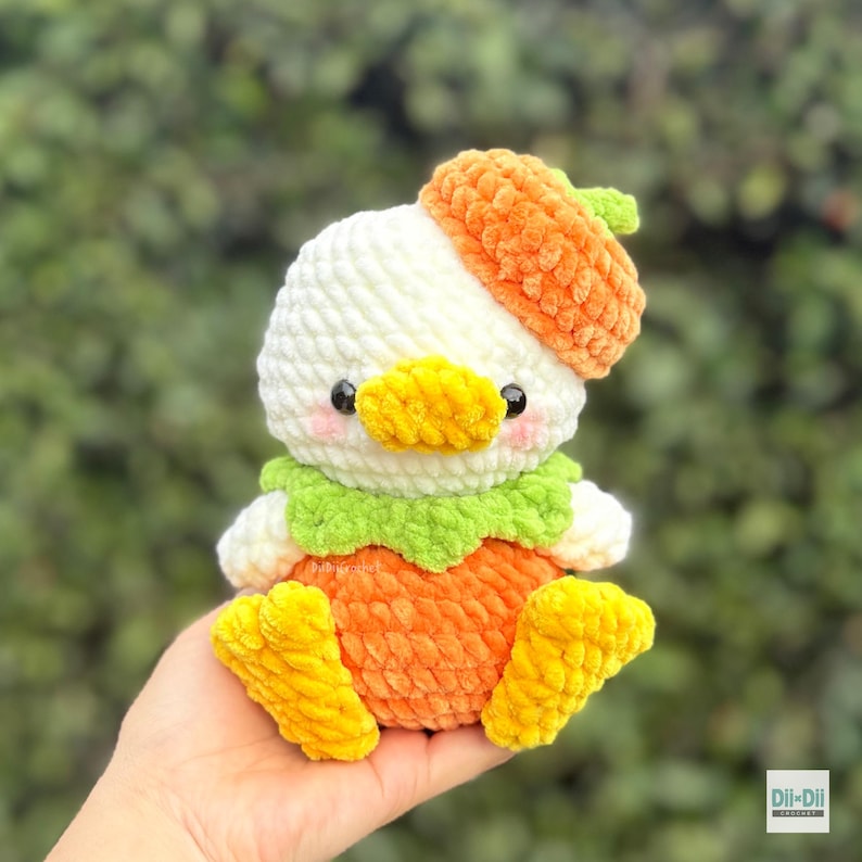 May include: A plush crochet toy shaped like a duck wearing an orange acorn cap. The duck has a white head, a yellow beak, pink cheeks, a green collar, and an orange body with yellow feet. The text Dil-Dil Crochet is visible in the bottom corner.