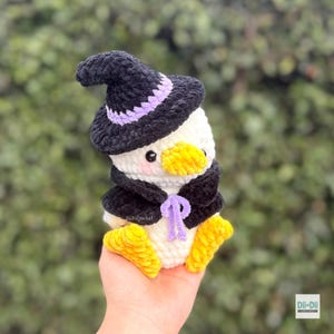 May include: A white crochet duck plushie wearing a black witch hat with a purple band and a matching black cape with a purple tie. The duck has a bright yellow beak and feet, black eyes, and pink blush on its cheeks. The text DiiDiiCrochet is visible.