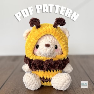 Könnte beinhalten: Ein weißer Plüschbär in einem gelb-braunen Bienenkostüm. Der Bär hat eine gelbe Kapuze mit braunen Fühlern, einen braunen Kragen und einen gelb-braun gestreiften Körper. Der Text "PDF PATTERN" steht über dem Bären.