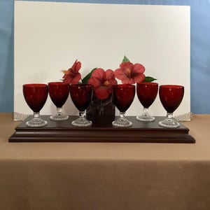 May include: Six ruby red wine glasses with clear, patterned stems are arranged on a dark wooden shelf. Red hibiscus flowers and greenery are behind the glasses. The glasses are set against a white backdrop.