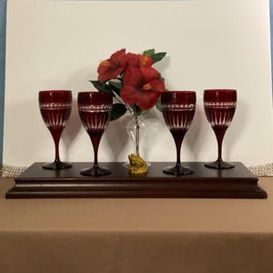 May include: Four ruby red wine glasses with etched designs are displayed on a dark wooden base. A small bouquet of red flowers and a decorative gold frog are also present. The glasses are approximately 7 inches tall.