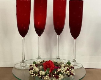 May include: Four red champagne flutes with clear stems and bases are arranged on a glass serving tray. A decorative wreath with white berries, red berries, and a red flower sits on the tray.