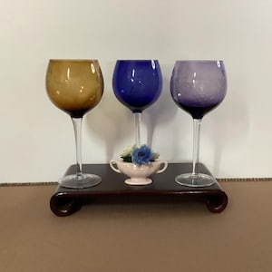 May include: Three colored wine glasses on a dark wooden stand. The glasses are amber, blue, and purple. A small pink dish with blue and white flowers sits on the stand. The glasses have long stems.