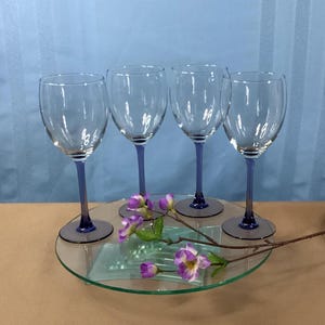 May include: Set of four clear glass wine glasses with blue stems and bases. The glasses are arranged on a clear glass tray with a sprig of purple flowers.