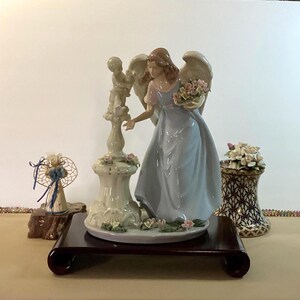 May include: A porcelain figurine of an angel with light brown hair and large wings, wearing a pale blue dress with pink sleeves. The angel holds a basket of pink and white flowers while standing next to a pedestal topped with a small cherub statue.