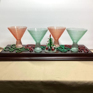 May include: Four stemmed cocktail glasses with a textured hobnail pattern, alternating between light pink and light green colors. A small glass Christmas tree figurine with red and green presents sits in the center among green and red beads.
