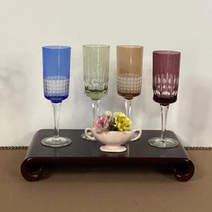 May include: Four colored champagne flutes in blue, green, amber, and purple, with a cut glass pattern on the lower half. They sit on a dark wood tray with a small pink dish of flowers.