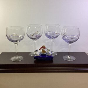May include: Four clear glass wine glasses with thin purple swirling lines painted around the bowls. Centered between the glasses is a small blue base featuring two white ceramic seal figurines and a miniature glass bottle with a cork stopper.