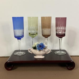 May include: Four colorful champagne flutes in blue, green, brown, and purple, with intricate cut glass designs. They stand on a dark wooden tray, accompanied by a small pink vase with blue and white flowers. A decorative display.