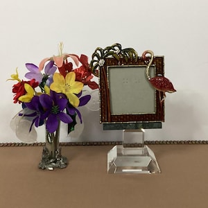 May include: A decorative picture frame with a red and brown border, featuring a flamingo design. The frame sits on a clear, geometric base. A vase of colorful artificial flowers is to the left.