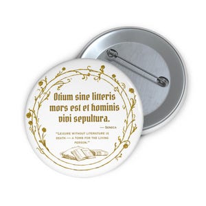 May include: White button with gold text and a gold floral border. The text reads "Otium sine litteris mors est et hominis vivi sepultura" and "Leisure without literature is death - a tomb for the living person." A second, silver button is partially visible.