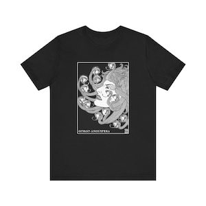 May include: Black t-shirt featuring a white illustration of Medusa with snakes for hair. The design is framed in a white rectangle, with the text "GORGO ANGUIFERA" below the image. The shirt is made of a soft material.