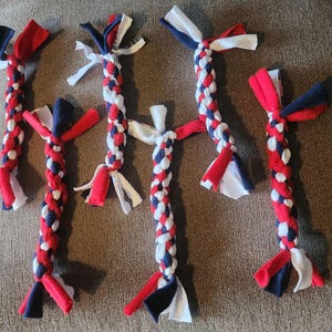 May include: Six braided fabric dog toys arranged in a group. Each toy features a repeating pattern of red, white, and navy blue fabric strips woven into a sturdy rope design with loose, knotted ends extending from both sides of the central braid.