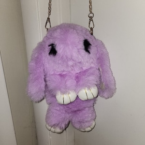 May include: A lavender plush bunny bag with a gold chain strap. The bunny has black eyes and white feet.