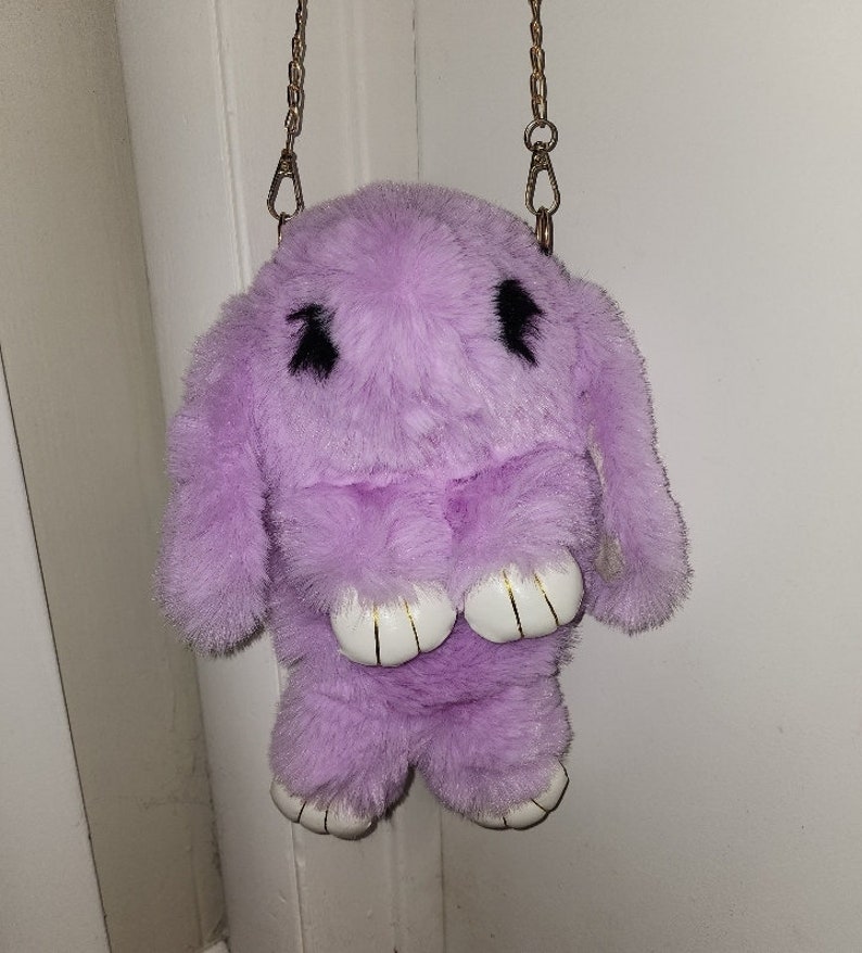 May include: A lavender plush bunny bag with a gold chain strap. The bunny has black eyes and white feet.