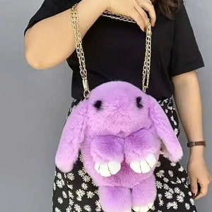 May include: A purple plush bunny shaped handbag with a gold chain strap. The bag is worn by a person with a black shirt and a floral skirt.