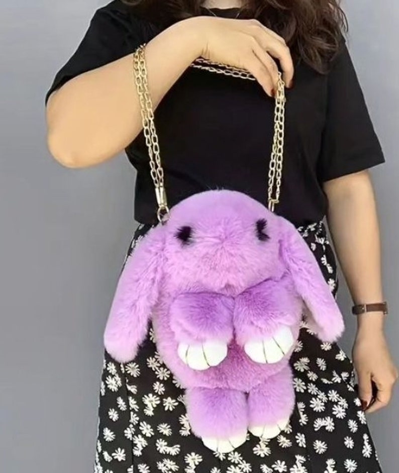 May include: A purple plush bunny shaped handbag with a gold chain strap. The bag is worn by a person with a black shirt and a floral skirt.