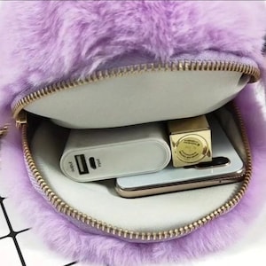 May include: A purple faux fur bag with a gold zipper. The bag is open and shows a white power bank, a gold lipstick, and a smartphone inside.