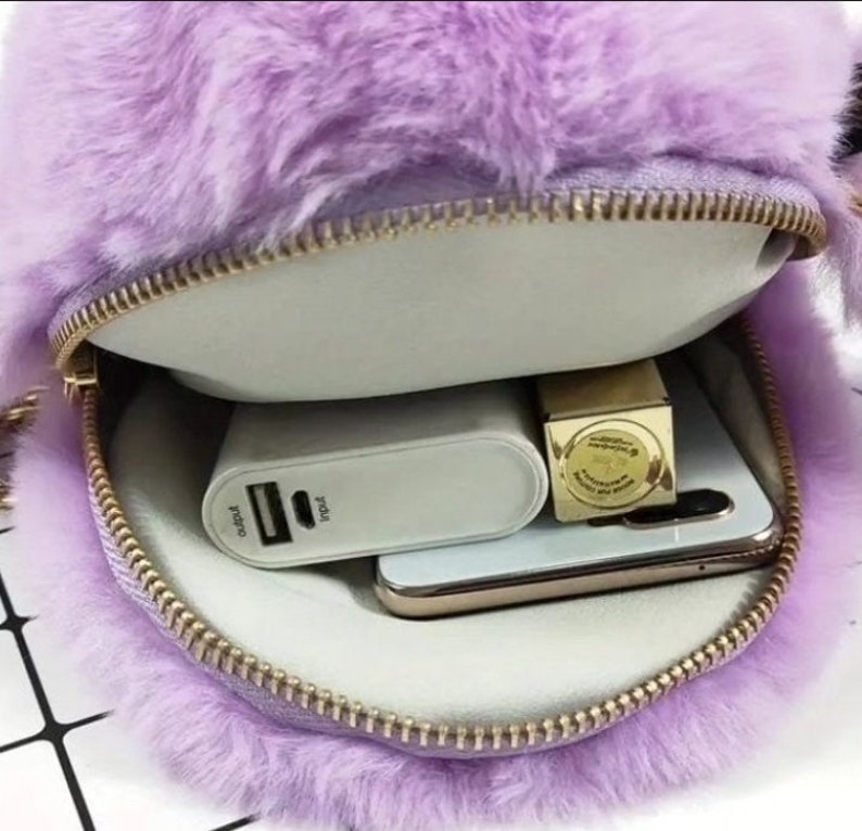 May include: A purple faux fur bag with a gold zipper. The bag is open and shows a white power bank, a gold lipstick, and a smartphone inside.