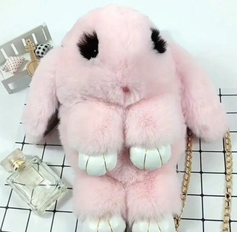 May include: A pink fluffy bunny shaped purse with a gold chain strap. The purse has black eyes and white paws.